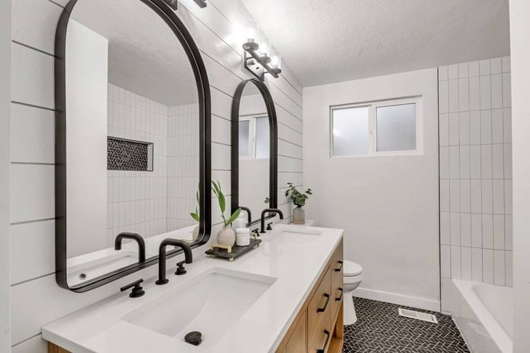 Designer bathroom with twin arched black-framed mirrors, light-wood double-sink vanity, vertical white subway tile, and hexagon-pattern black floor — Utah bathroom remodel by CasaMás Construction